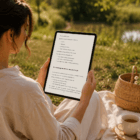 Osoba siedząca na kocu w otoczeniu natury czyta ebook „Reset układu nerwowego – 21 dni” na tablecie, obok kubka herbaty, notesu i kosza z kwiatami.